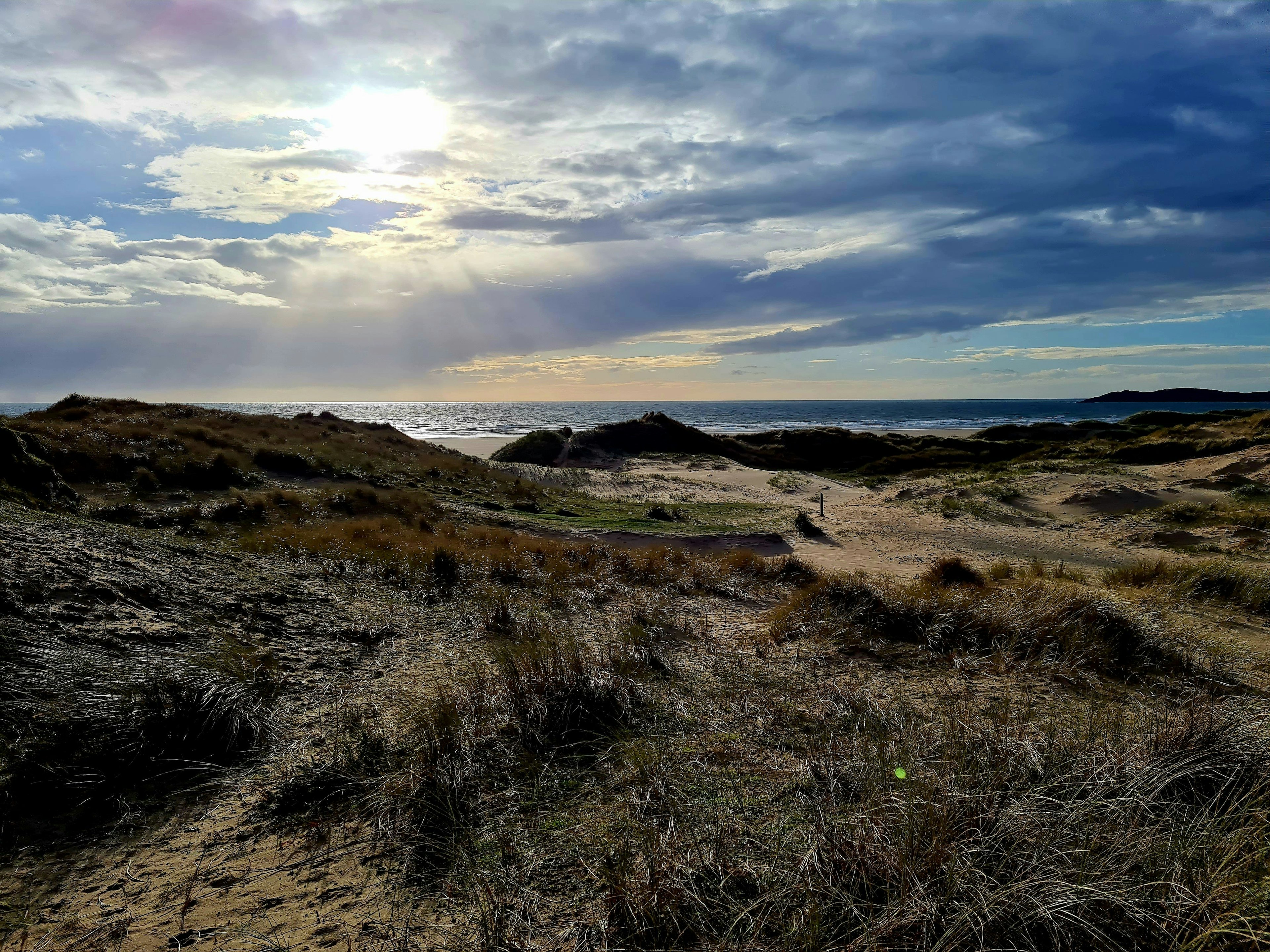 Sand dunes and grass with the ocean on the horizon and sun beams coming through the clouds