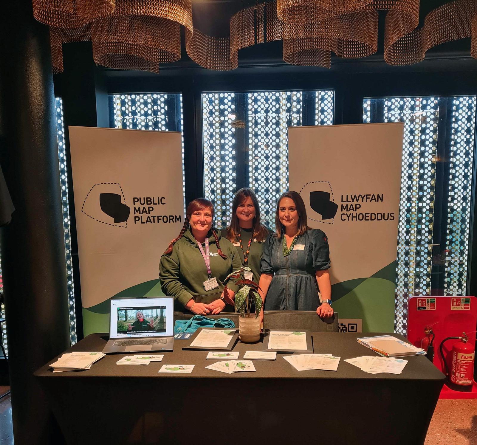 Three women from the Public Map Platform team stand proudly at a PMP event stand