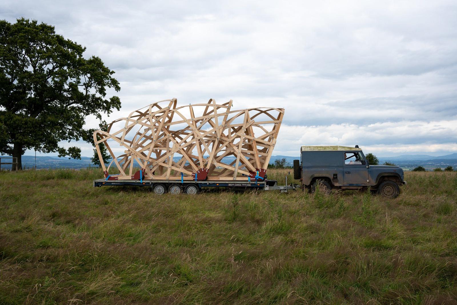 Amddiffynnwr Landrover glas gyda threlar ac arno set o strwythurau pensaernïol diddorol wedi'u gwneud o bren wedi'i blygu.