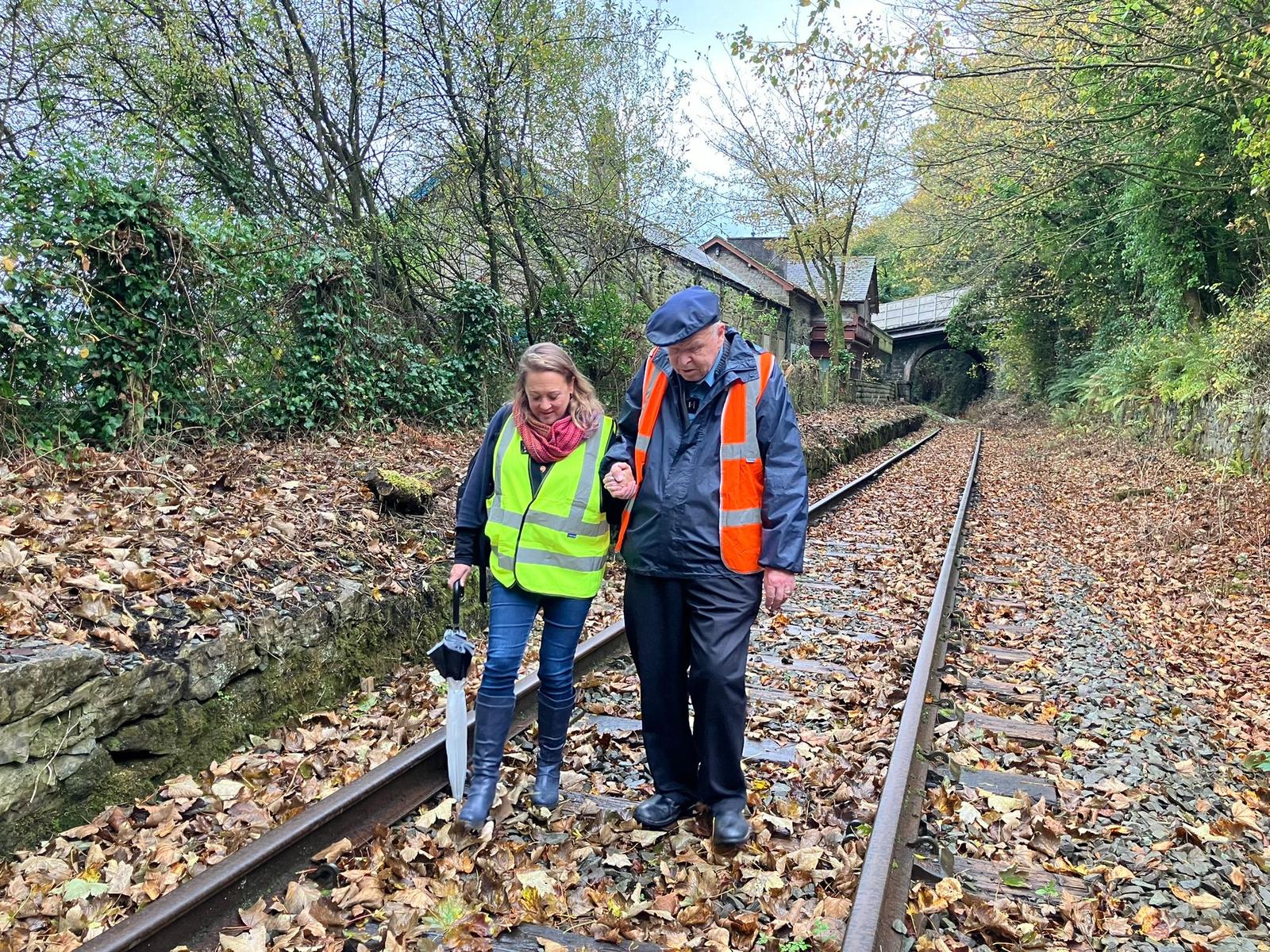 Gŵr bonheddig hŷn a dynes yn cerdded braich-yn-braich ar hyd trac rheilffordd segur. Mae'r ddau yn gwisgo festiau hi-viz ac mae'r fenyw yn cario ambarél. Mae dail yr hydref wedi cwympo o'u cwmpas ac mae'r grŵp yn llaith.