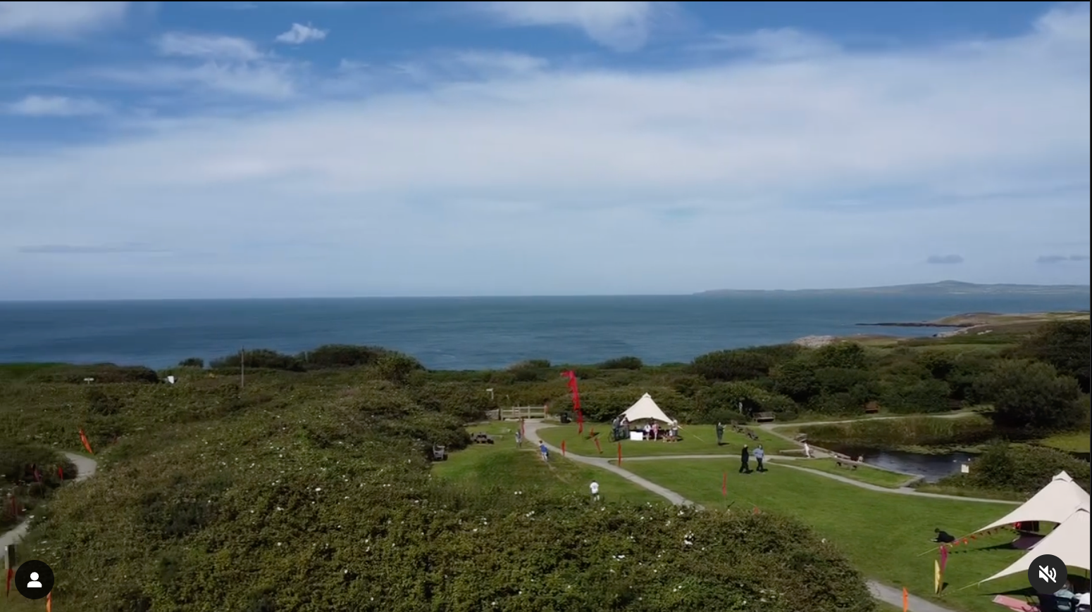 A drones eye view of the sea foregrounded by a few tent canopy structures set in a green and bushy landscape