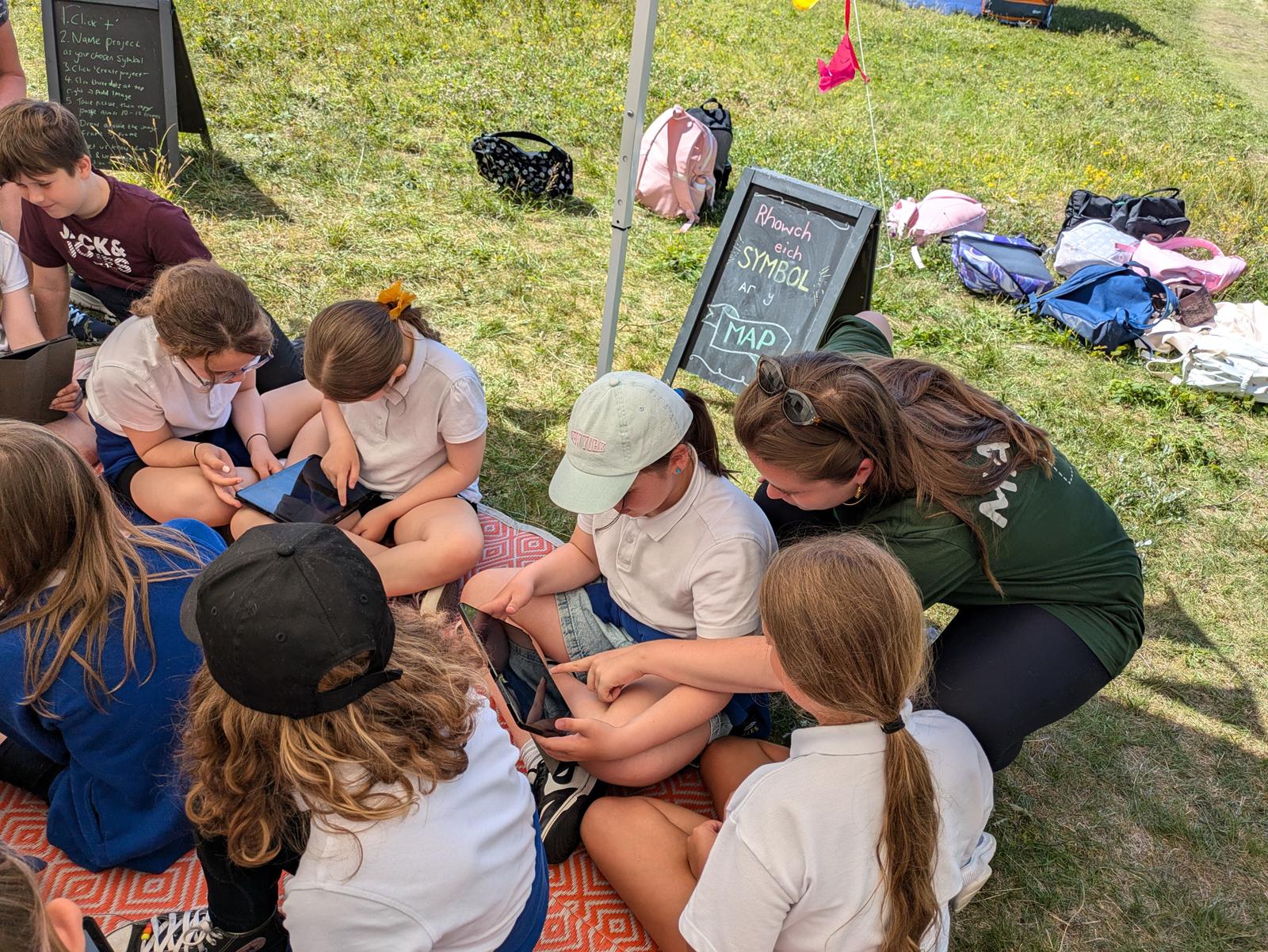 A group of schoolchildren are sat in the shade outside, using tablets to make digital animations. A member of staff helps one of them with their drawing.