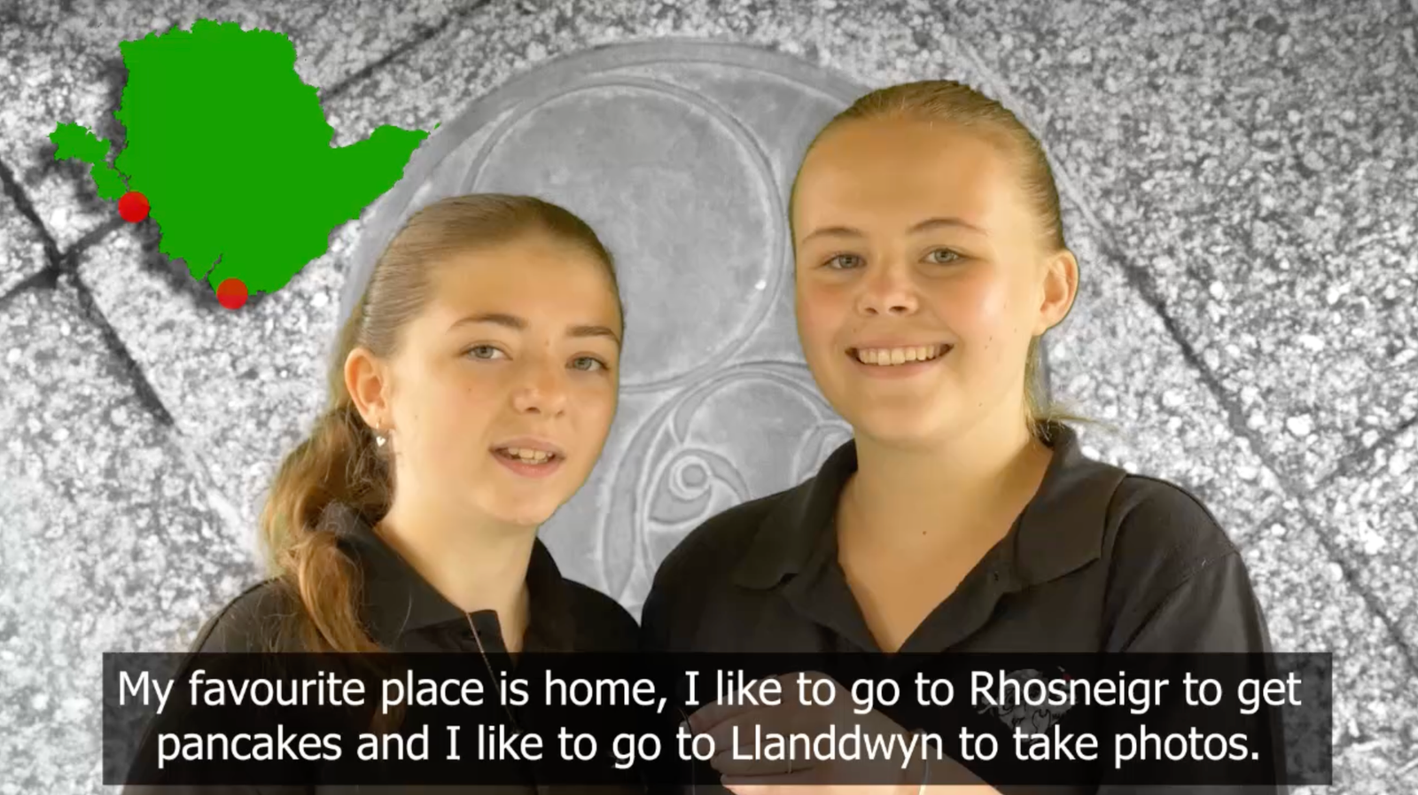 Two young girls in black polo shirts are stood side by side, smiling. In the top left corner is a map of Anglesey with two location markers on them. The image features a caption of the girls speech reading: 'My favourite place is home, I like to go to Rhosneigr to get pancakes and I like to go to Llanddwyn to take photos."