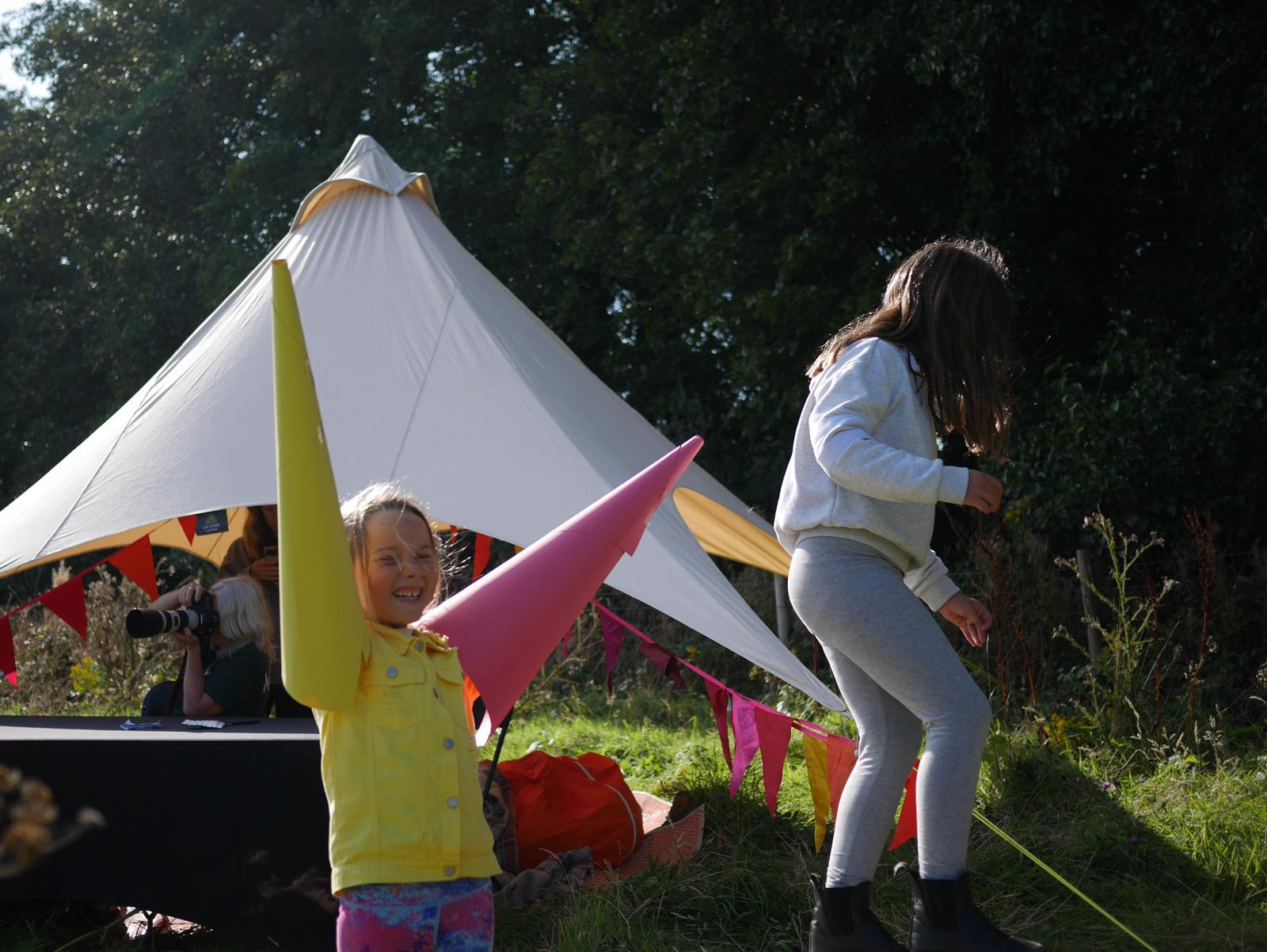 A young girl in a yellow jacket has put a listening cone on either arm.